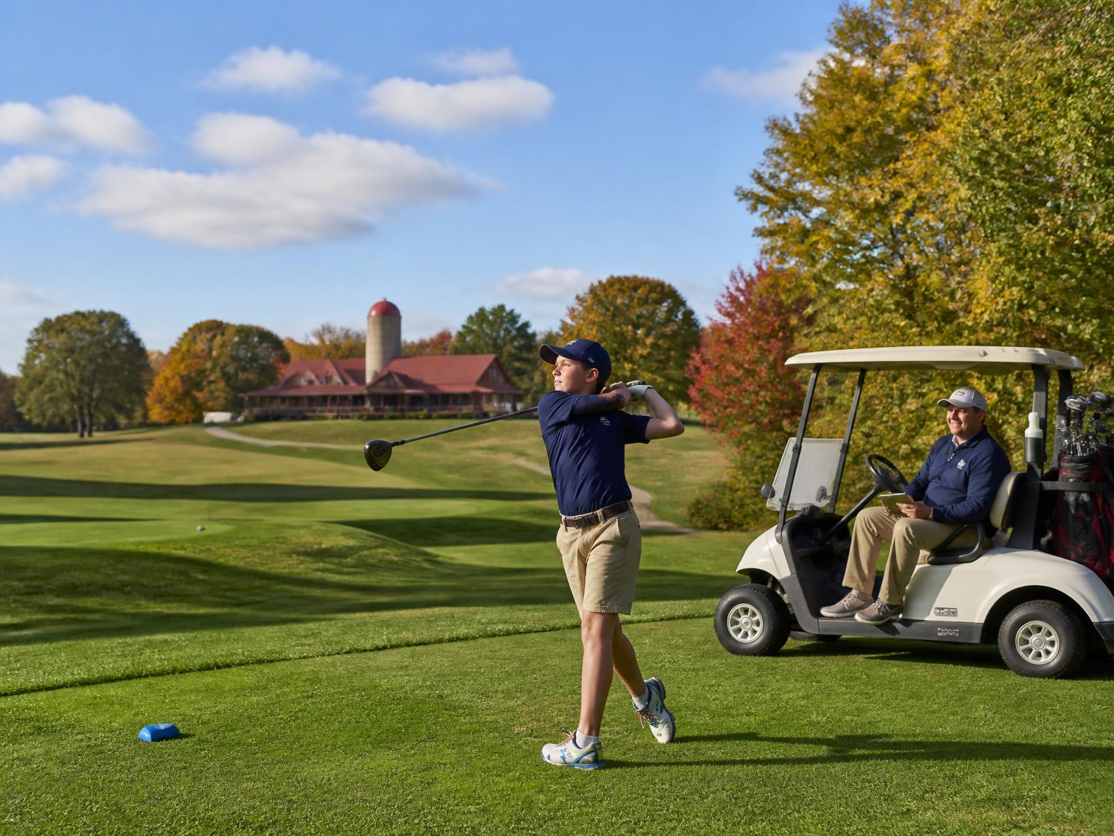 A middle school golfer teeing off on a sunny early fall afternoon at a scenic golf course while a parent watches from a cart near the clubhouse